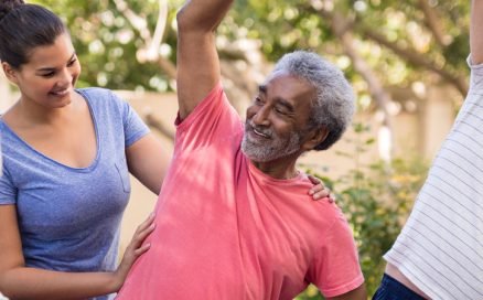 An older man stretching one arm overhead outdoors while a wellness instructor supports him.