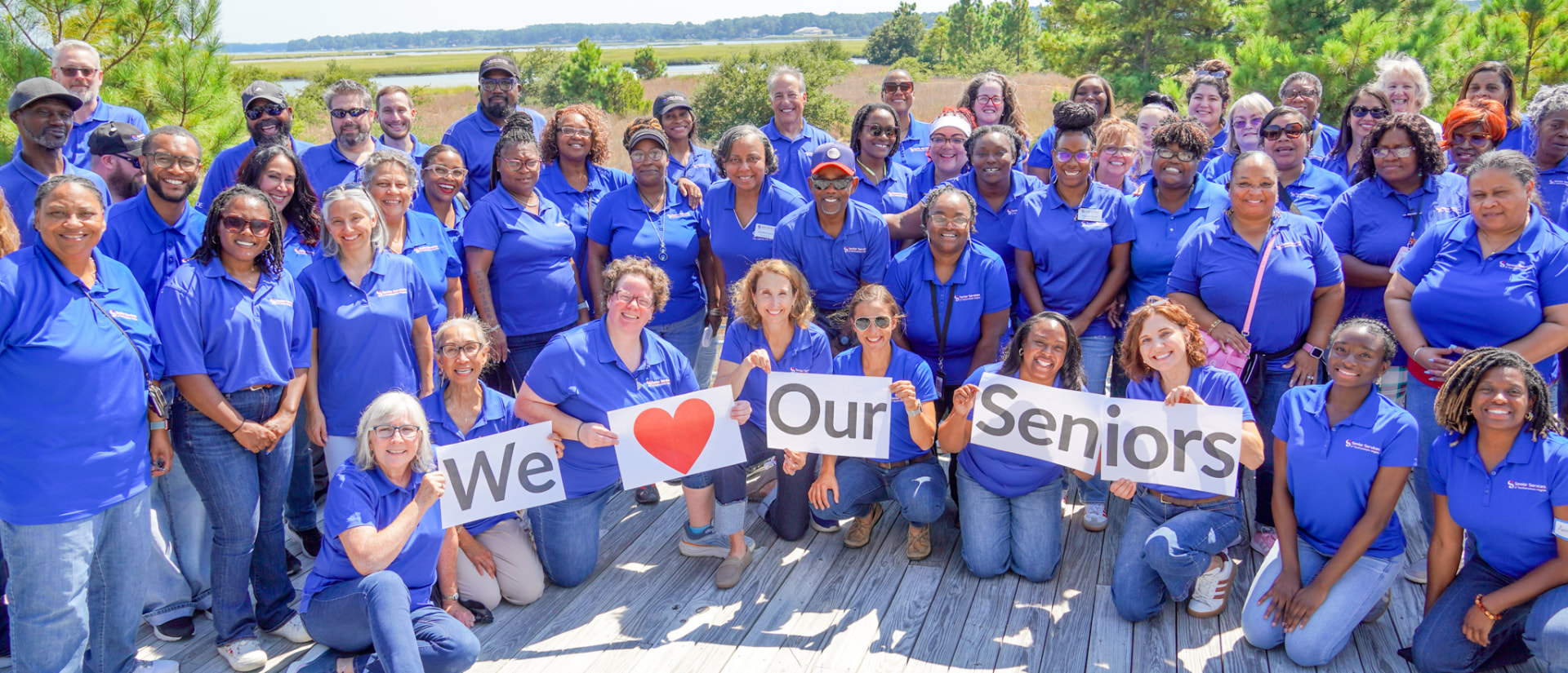 The Senior Services staff team gathered on a marsh boardwalk holding signs that read “We love our seniors”.