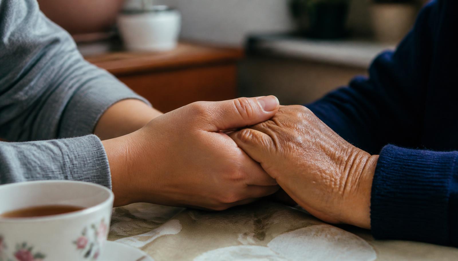 A younger hand and an elderly hand clasped together across a kitchen table.